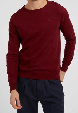 Mann trägt einen eng anliegenden dunkelroten Pullover mit Rundhalsausschnitt und weite dunkelblaue plissierte Hose vor einem schlichten Hintergrund.