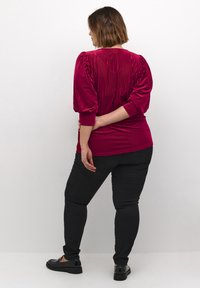 Top rouge à côtes avec des manches bouffantes, associé à un jean noir ajusté et des chaussures noires, vu de dos sur un fond neutre.