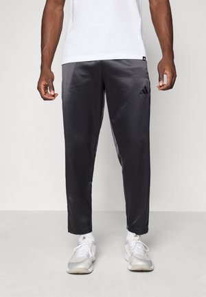 Homme portant un pantalon de survêtement Adidas noir, des chaussettes blanches et des baskets blanches Adidas, debout devant un fond clair uni.