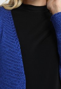 Modrý pletený cardigan s otevřeným předním dílem, s texturovaným pletením, nošený přes černý top s vysokým límcem.