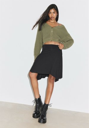Femme portant un pull court boutonné vert olive, une jupe asymétrique noire et des bottines à plateforme noires lacées, debout contre un mur blanc.
