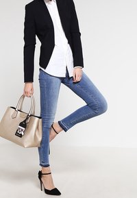 Svart kavaj över en vit skjorta, blå skinny jeans, svarta högklackade skor, håller en metallisk gyllene handväska med en tag.