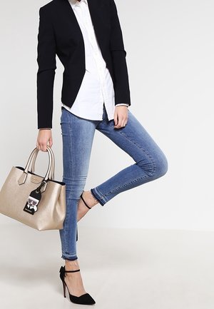 Blazer noir sur une chemise blanche, jean skinny bleu, chaussures à talons noirs, tenant un sac à main doré métallique avec une étiquette.