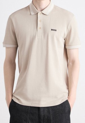 Beiges Polohemd mit kurzen Ärmeln, Kragen und Knöpfen, mit dem "BOSS"-Logo auf der linken Brust, getragen mit dunkler Hose.
