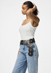 Zwarte leren riem met ronde accenten en een decoratieve zilveren gesp, gedragen over hoge blauwe spijkerbroeken en een witte tanktop.
