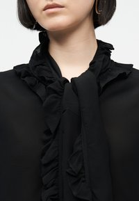 Mujer con cabello corto y oscuro, vistiendo una blusa negra con cuello de volantes atado en un lazo y pendientes de aro dorados, vista desde la barbilla hasta el pecho.