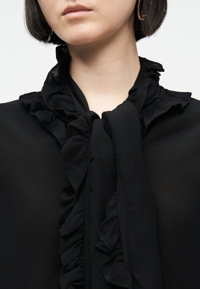 Γυναίκα με κοντά σκούρα μαλλιά που φοράει μια μαύρη μπλούζα με βολάν στο γιακά δεμένο φιόγκο και χρυσούς κρίκους στα αυτιά, φαίνεται από το πηγούνι μέχρι το στήθος.