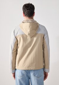Beige og lysegrå vindjakke med hette, med en lett tekstur og kontrasterende skulderpaneler.