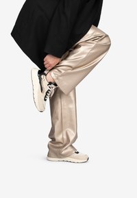 Beige, lederähnliche Hosen und eine übergroße schwarze Jacke, getragen mit Sneakers, die ein strukturiertes cremefarbenes und schwarzes Design aufweisen.