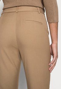 Pantalon beige sur mesure avec une texture lisse, doté d'une poche arrière et d'une silhouette ajustée, associé à un pull côtelé couleur noisette.