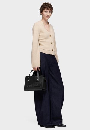 Donna che indossa un cardigan beige con bottoni e pantaloni larghi scuri, che tiene una borsa nera con doppi manici.