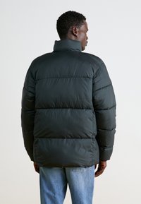 Sort pufferjakke med høj krave, quiltet design og ribbede manchetter. Båret over lyseblå jeans. Enkel, struktureret stof med en glat finish.