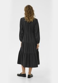 Woman with wavy blonde hair wearing a long-sleeve, black tiered midi dress and black loafers, standing facing away on a white background.