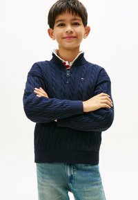 Tumši zils kabelisadītā džemperis ar rāvējslēdzēja apkakli un ribotām piedurknēm. Kombinēts ar gaiši ziliem džinsiem. Tīrs, minimālisma fons.