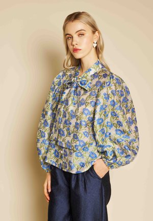 Femme portant un chemisier à fleurs bleues avec des manches bouffantes, associé à un pantalon sombre texturé, debout devant un fond beige.