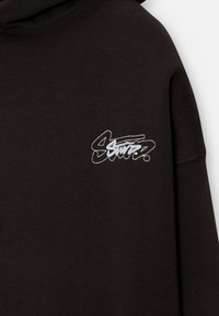 Czarna bluza z kapturem z białym, haftowanym logo na lewej piersi. Materiał jest miękki i teksturowany, z klasycznym kapturem.