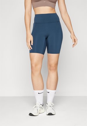 Kobieta w ciemnoniebieskich, wysokich szortach rowerowych, białych skarpetkach Nike, białych trampkach i brązowym sportowym biustonoszu, stojąca na jednolitym tle.
