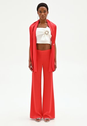 Donna che indossa pantaloni rossi a gamba larga e vita alta, crop top bianco con fibbia decorativa, e giacca rossa appoggiata sulle spalle, in piedi dritta.