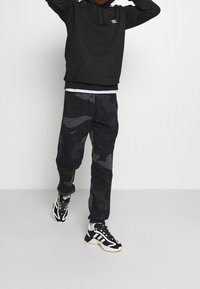 Schwarzer Hoodie mit einer Vordertasche, graue Camouflage-Sweatpants mit blauen Flecken und schwarz-weiße Laufschuhe. Minimalistisches Design und bequeme Passform.