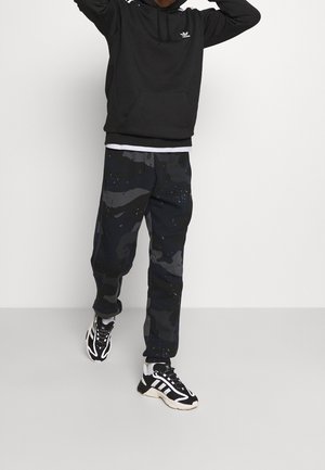 Schwarzer Hoodie mit einer Vordertasche, graue Camouflage-Sweatpants mit blauen Flecken und schwarz-weiße Laufschuhe. Minimalistisches Design und bequeme Passform.