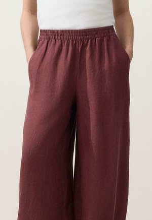 Donna che indossa pantaloni bordeaux larghi a vita alta e gamba larga con elastico in vita e mani nelle tasche, abbinati a una maglietta bianca.