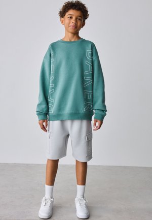 Garçon aux cheveux bouclés portant un sweat-shirt oversize bleu sarcelle avec l'inscription "DAXER" et un short cargo gris clair avec des baskets blanches.