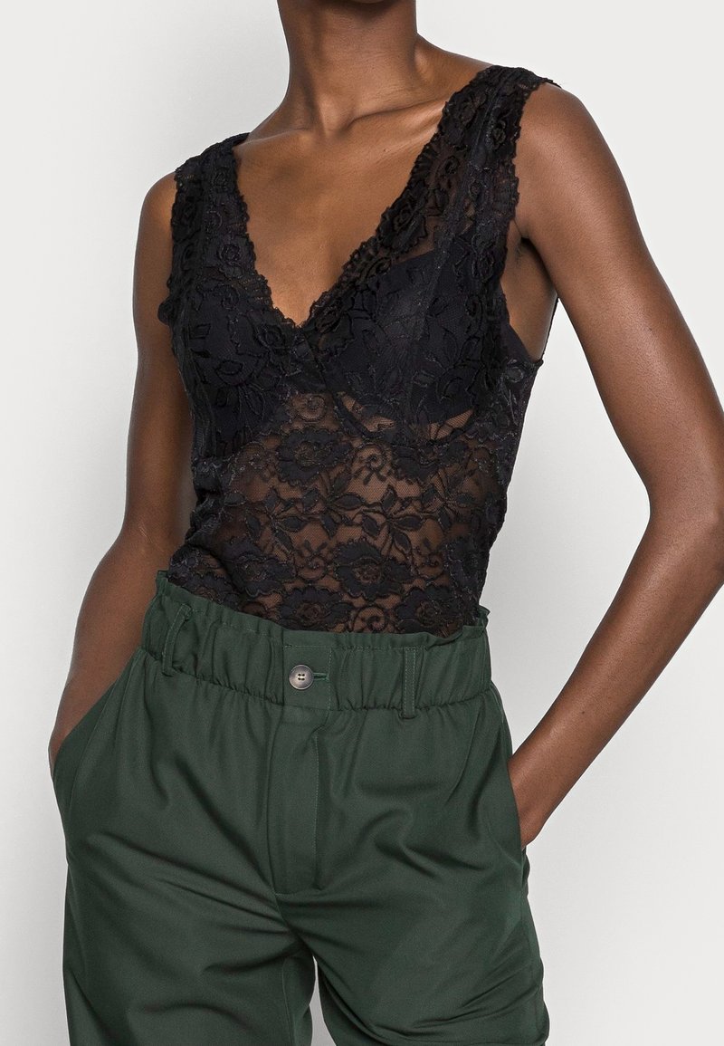 Bodysuit en dentelle noire avec un décolleté en V profond, motif floral et tissu transparent, associé à un pantalon taille haute vert avec une taille froncée.