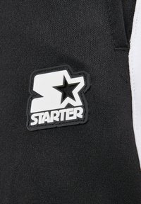 Melnā audumā ar smalku tekstūru ir balts un melns gumijas logotipa pleķis. Šajā pleķī ir vārds "STARTER" un zvaigznītes dizains.