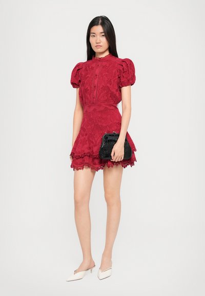 Rood bloemenpatroon jurk met opbollende mouwen, kantafwerking en een strakke taille, gecombineerd met een zwarte tas en crèmekleurige sleehakken.