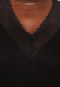 Černý pletený oděv s výstřihem do V zdobeným složitou černou krajkou, hladkou texturou a jemnými žebrovými detaily.