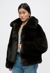 Καφέ ψεύτικο παλτό από γούνα με μεγάλο γιακά, μαλακή υφή και χαλαρή εφαρμογή; συνδυασμένο με ανοιχτόχρωμο μπλουζάκι και μπλε τζιν.