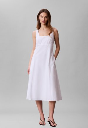 Kobieta nosząca białą, bezrękawną sukienkę midi z kwadratowym dekoltem i kieszeniami, w połączeniu z czarnymi sandałami na obcasie z paskami, stojąca na szarym tle.