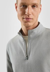 Młody mężczyzna ubrany w jasnoszary sweter z prążkowanej tkaniny z zamkiem błyskawicznym do jednej czwartej oraz srebrnymi kolczykami w kształcie kółek na jednolitym tle.