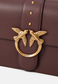 Maroon leather handbag with embossed birds and a circular gold-toned hardware closure. Decorative studs enhance the surface texture.