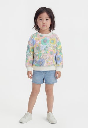 Dieťa oblečené v pastelovej kvetinovej mikine, rifľových krátkych nohaviciach a bielych teniskách, stojace pred jednoduchým bielym pozadím.