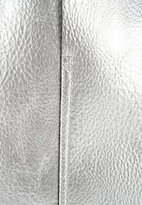 Zilveren metallic leer met een gestructureerde afwerking. Heeft een centrale naad gestikt met witte draad, wat de glans van het materiaal benadrukt.
