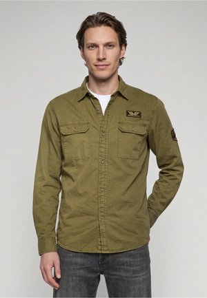 Homme portant une chemise boutonnée vert olive avec poches poitrine et écussons, debout devant un fond clair uni.