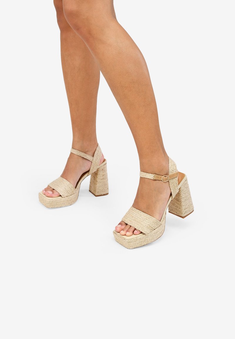 Sandalias de plataforma beige con correas anchas trenzadas y un tacón grueso y texturizado. Cinta ajustable en el tobillo con hebilla. Diseño de punta abierta.