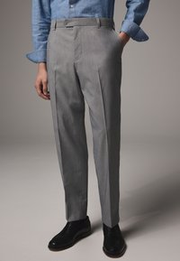 Pantaloni grigi da donna con vestibilità sartoriale, caratterizzati da due pieghe frontali, tasche laterali e chiusura con bottone, abbinati a scarpe nere.