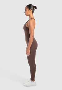 Bruine leggings met een strakke pasvorm, gemaakt van rekbaar materiaal, gedragen met witte sportschoenen. Het zijprofiel toont de naadloze constructie.