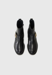 Botas de tornozelo em couro preto com uma textura suave, apresentando um fecho lateral de correr e um detalhe em fivela dourada. Bico arredondado e sola plana.