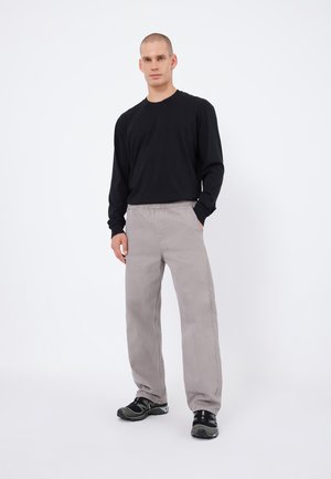 Cămașă neagră cu mânecă lungă, pantaloni gri deschis cu un elastic în talie și adidași negri cu accente verzi, stând pe un fundal alb.