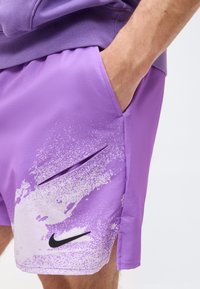 Άνδρας φοράει μωβ σορτς Nike με λευκή σχεδίαση πιτσιλιών μπογιάς και μαύρο λογότυπο swoosh, χέρι στην πλαϊνή τσέπη σε λευκό φόντο.