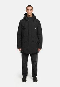 Čierna parka s kapucňou, zapínanie na patent a zips, dve predné vrecká a nastaviteľné sťahovacie šnúrky. Nosená cez tmavo textúrované nohavice a topánky.