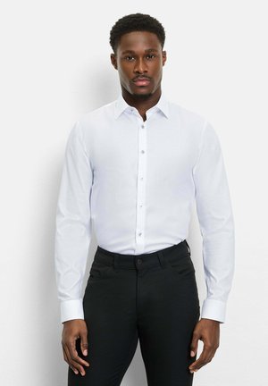 Mann trägt ein eng anliegendes weißes Hemd mit langen Ärmeln und schwarzen Hosen, steht vor einem schlichten weißen Hintergrund.