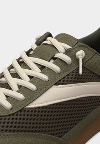 Κοντινό πλάνο ελιάς πράσινου αθλητικού παπουτσιού με λευκά κορδόνια, πλευρές από δίχτυ, σουέτ λεπτομέρειες και λευκή διακριτική ρίγα πάνω σε σόλα καραμέλας.