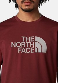 Camiseta de algodón color burdeos con un gran diseño de logotipo blanco de "The North Face" en el pecho. Cuello redondo y mangas cortas.