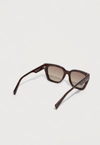 Γυαλιά ηλίου χελώνας με υπερμεγέθη, γωνιώδη πλαίσια και καφέ φακούς με διαβάθμιση χρώματος. Διαθέτουν υφασμένα μπράτσα με έγχρωμες λεπτομέρειες.