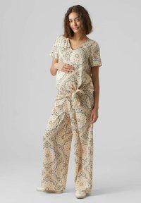 Outfit premaman con una maglietta crema a maniche corte e un dettaglio nodo, abbinata a pantaloni larghi a stampa in morbidi colori pastello.