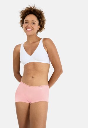 Smilende kvinde med krøllet hår iført en hvid bh og lyserøde boyshorts, stående med hænderne bag ryggen foran en hvid baggrund.